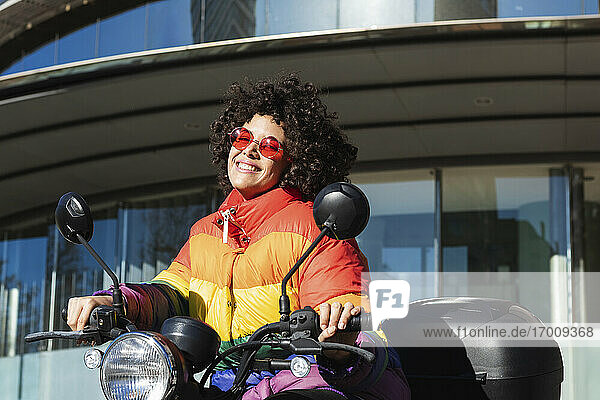 Frau mit Sonnenbrille und bunter Jacke lächelnd auf einem Motorrad sitzend
