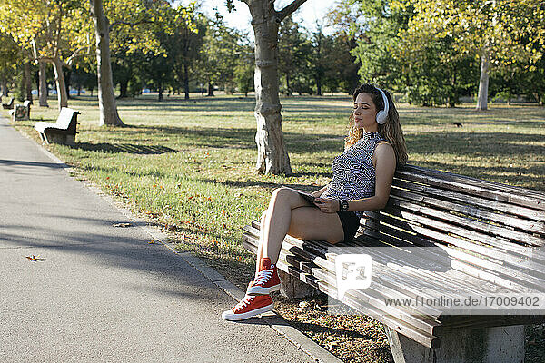 Junge Frau mit geschlossenen Augen hört Musik über Kopfhörer in einem öffentlichen Park an einem sonnigen Tag