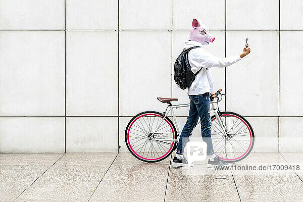 Young man with bicycle wearing pig mask while taking selfie on mobile phone