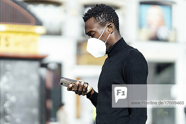 Junger Mann mit Gesichtsmaske  der im Freien stehend ein Mobiltelefon benutzt