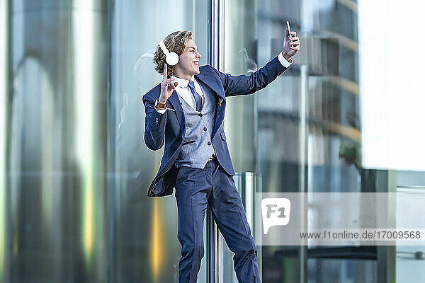 Glückliche männliche professionelle Geste bei der Aufnahme von Selfie durch Smartphone gegen Bürogebäude