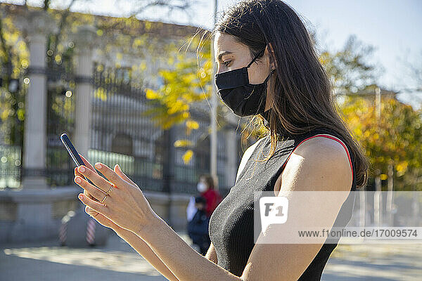 Junge Frau mit Gesichtsmaske  die ein Smartphone benutzt  während sie auf der Straße steht
