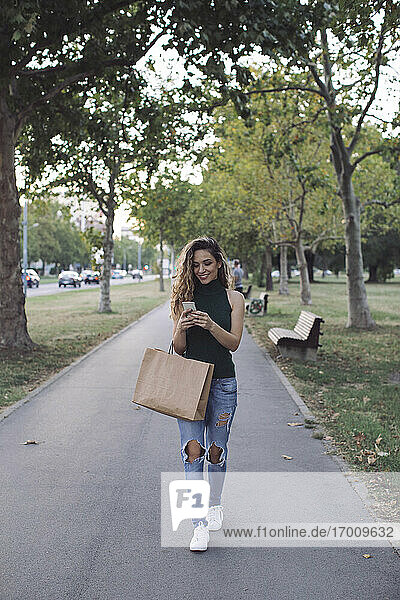 Glückliche junge Frau mit Papiertüte und Smartphone beim Spaziergang im Park