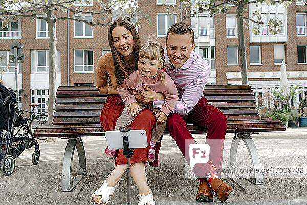 Glückliche Familie mit einer Tochter  die ein Selfie mit ihrem Handy auf einem Stativ auf einer Parkbank macht