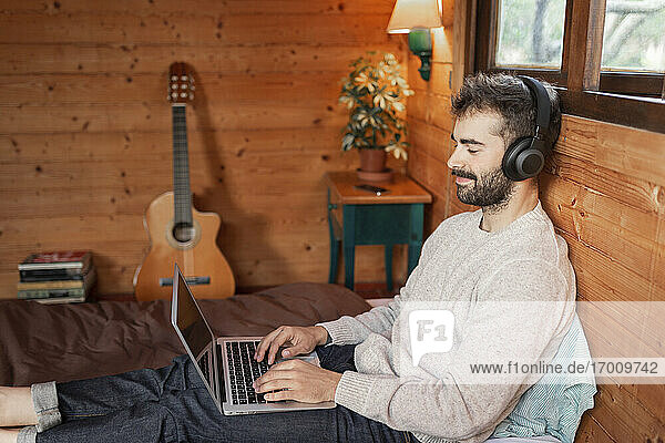 Lächelnder junger Mann  der einen Laptop benutzt und auf einem Bett in einer Blockhütte Musik hört