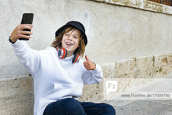 Boy wearing hat and headphones using mobile phone while sitting against wall