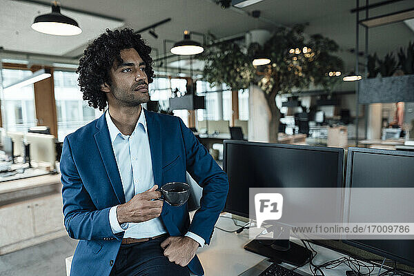 Nachdenklicher männlicher Unternehmer  der eine Kaffeetasse in der Hand hält  während er neben Computermonitoren am Schreibtisch im Büro sitzt