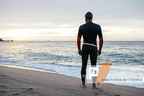 Männlicher Schwimmer  der allein am sandigen Küstenstrand steht  mit Schwimmflügel in der Hand