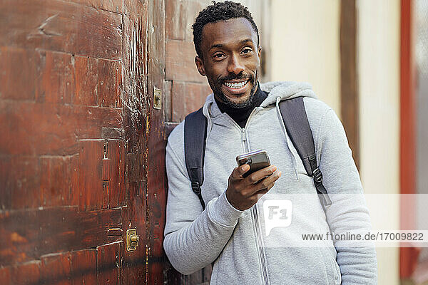 Man with backpack and mobile phone smiling while leaning on wall