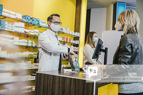 Weiblicher Kunde mit Gesichtsmaske beim Bezahlen mit dem Smartphone an der Kasse in einer Apotheke