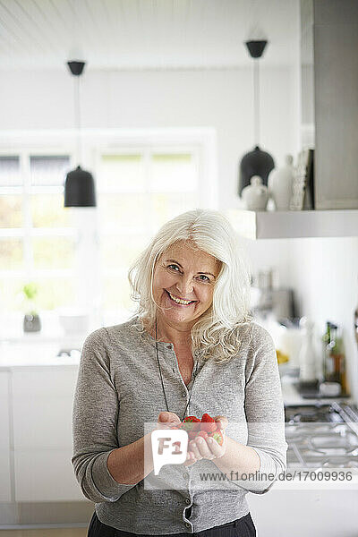 Smiling retired woman holding fresh strawberries while standing in kitchen at home