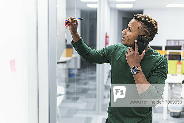 Male professional on phone call writing in adhesive note at work place