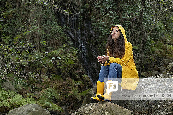 Nachdenklicher Wanderer im Regenmantel auf einem Felsen im Wald sitzend