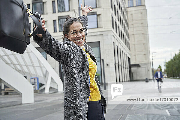Cheerful businesswoman holding laptop bag while standing with arms raised in city