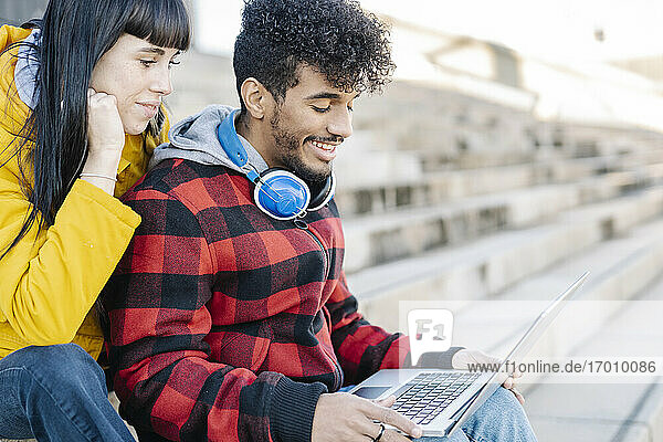 Junges Paar lächelt  während es im Freien sitzend einen Laptop benutzt