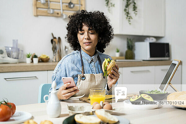 Porträt einer jungen Frau  die am Küchentisch mit einem Sandwich in der Hand ein Smartphone benutzt