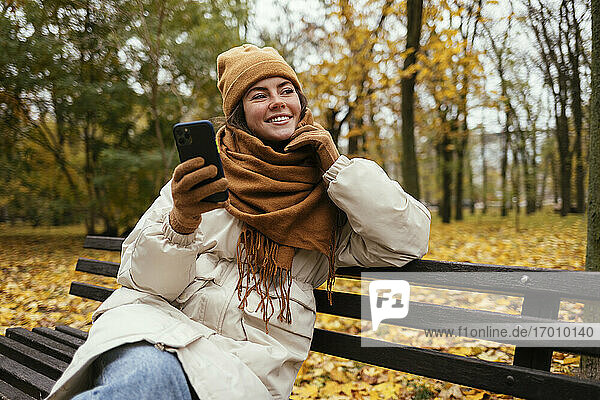 Lächelnde Frau  die im Herbst in einem Park ihr Handy in der Hand hält und träumt