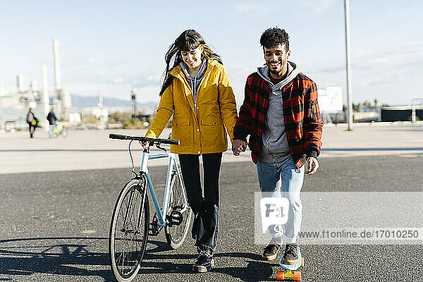 Smiling man skateboarding while holding hand of friend walking with bicycle on road
