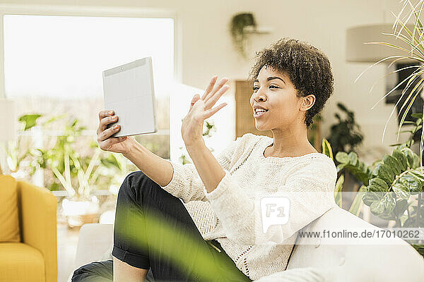 Frau winkt mit der Hand zu einem Videoanruf auf einem digitalen Tablet  während sie zu Hause sitzt