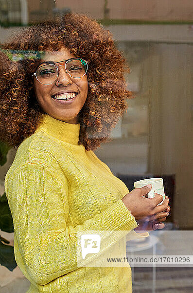 Lächelnde Frau mit Kaffeetasse träumt hinter Glas in einem Cafe