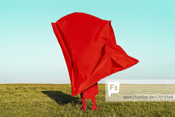 Woman covering herself with red fabric while standing on field against blue sky