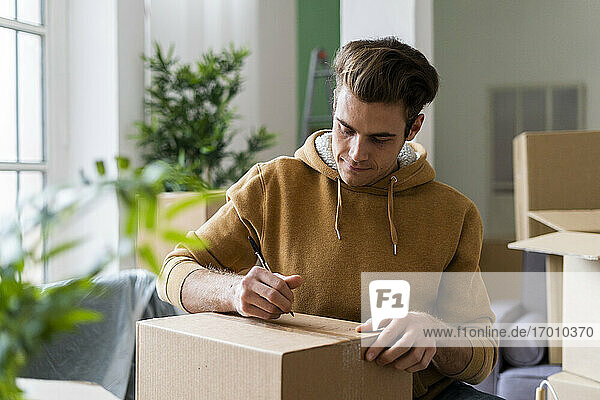 Junger Mann schreibt mit Stift auf Karton im neuen Zuhause