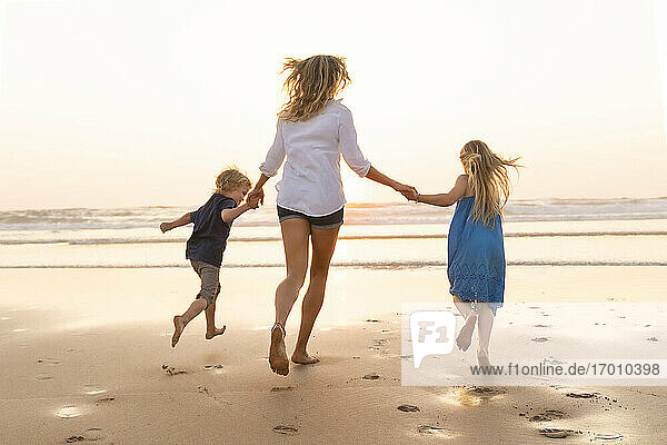 Kinder halten die Hände der Mutter beim Laufen am Strand während des Sonnenuntergangs