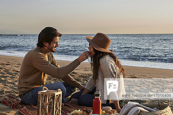 Smiling boyfriend feeding food to girlfriend while sitting on beach