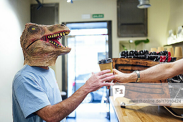 Hand eines Barista  der einem Mann mit Dinosauriermaske in einem Café Kaffee serviert