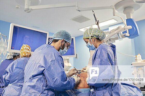 Ärzte mit Gesichtsmaske  die eine Operation durchführen  während sie mit einem Kollegen im Hintergrund im Operationssaal stehen