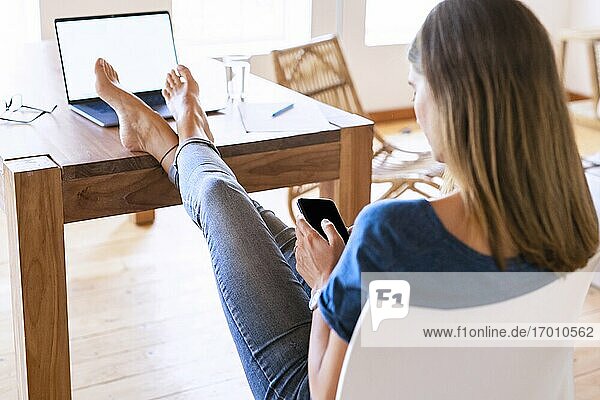 Brünette Geschäftsfrau, die ihr Smartphone benutzt, während sie sich mit hochgelegten Füßen am Tisch im Heimbüro entspannt