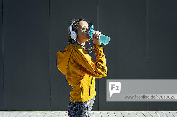 Weibliche Sportlerin mit Kopfhörern  die Wasser trinkt  während sie an einer grauen Wand steht