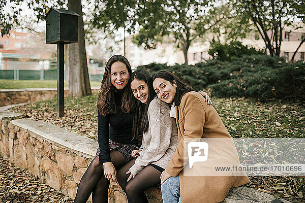 Lächelnde reife Frau mit Töchtern  die auf einer Stützmauer im Park sitzen