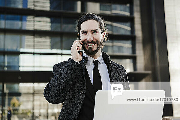 Lächelnder Geschäftsmann mit Laptop  der im Freien sitzend mit seinem Mobiltelefon spricht