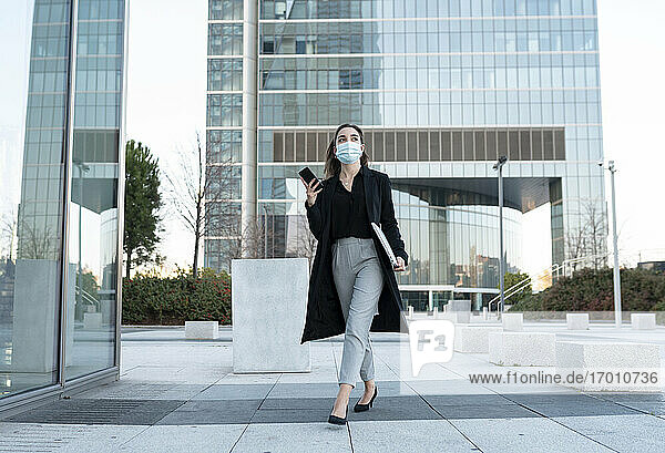 Geschäftsfrau mit Gesichtsmaske  die ein Mobiltelefon benutzt  während sie auf dem Gehweg geht