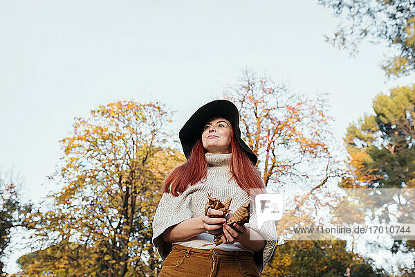 Thoughtful woman wearing hat holding autumn leaves while standing against clear sky in park
