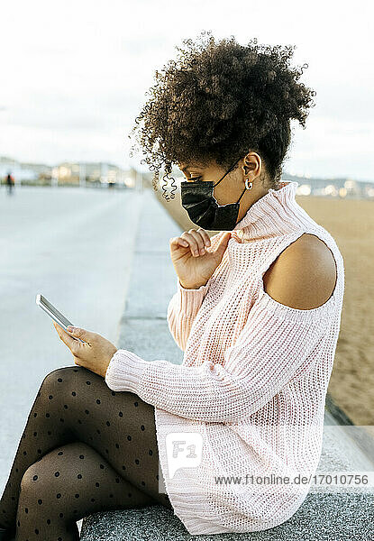 Frau mit lockigem Haar  die eine Gesichtsmaske trägt und ein Smartphone benutzt  während sie auf einer Stützmauer sitzt
