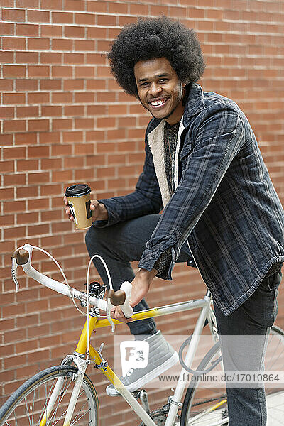Smiling young man holding coffee while sitting on bicycle against brick wall