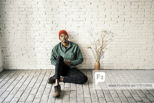 Mid erwachsenen Mann mit leeren Ausdruck sitzt von getrockneten Pflanze Vase gegen weiße Backsteinmauer