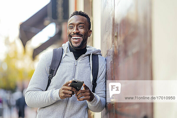 Lächelnder Mann  der ein Mobiltelefon benutzt und sich an die Wand lehnt