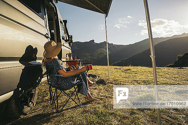 Frau auf Stuhl sitzend mit Kaffeetasse am Wohnmobil an einem sonnigen Tag