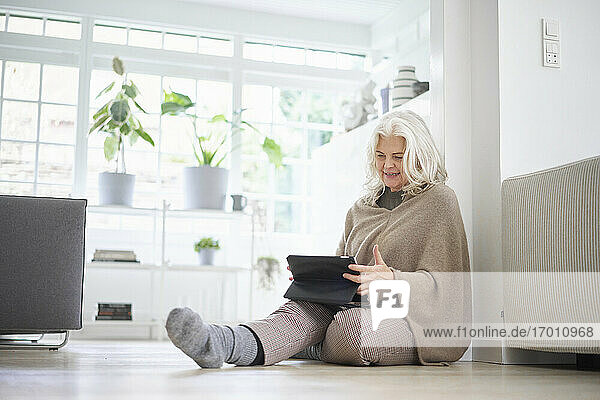 Ältere Frau im Ruhestand sitzt auf dem Boden und benutzt ein digitales Tablet im Wohnzimmer einer Wohnung