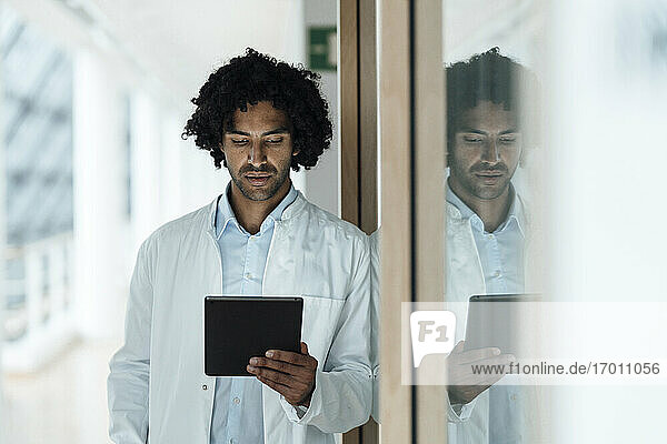 Young male doctor using digital tablet while leaning on glass wall at hospital
