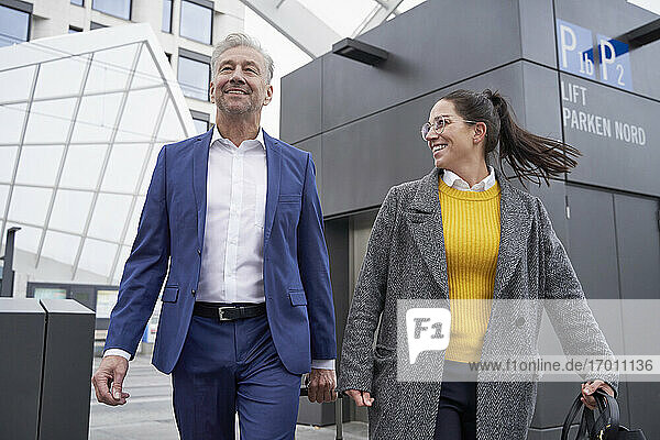 Lächelnde Geschäftsfrau und Geschäftsmann im Gespräch beim Gehen gegen Aufzug an der Bushaltestelle in der Stadt