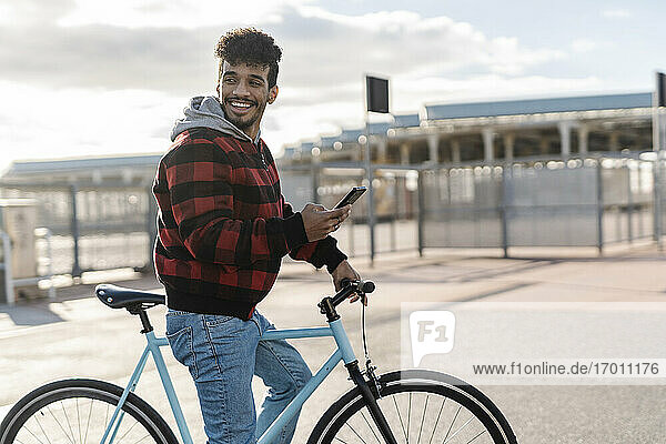 Lächelnder Mann mit Mobiltelefon  der wegschaut  während er an einem sonnigen Tag auf dem Fahrrad steht