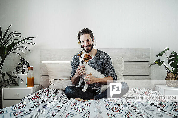 Hübscher Mann sitzt mit Hund auf dem Bett zu Hause