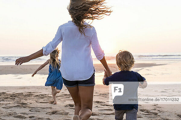 Mutter hält die Hand ihres Sohnes  während sie mit ihrer Tochter im Hintergrund am Strand läuft