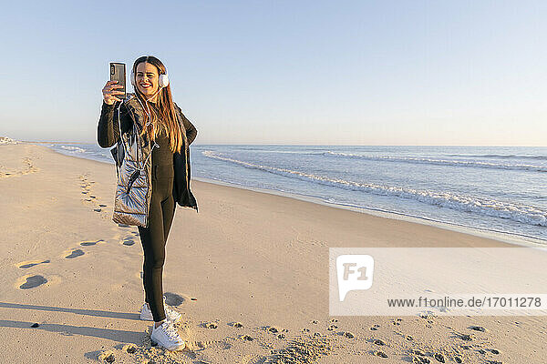 Smiling woman taking selfie with smart phone at beach against clear sky