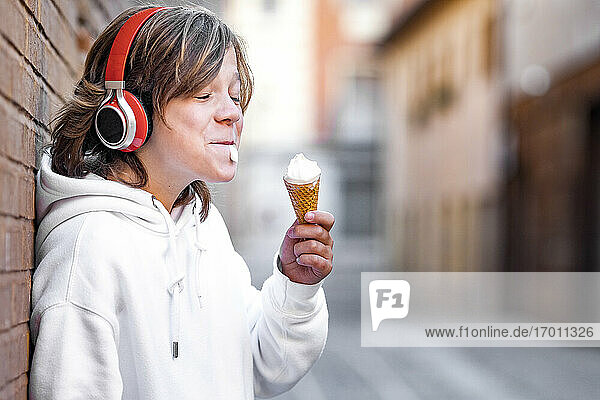 Boy wearing headphones eating ice cream while leaning on wall