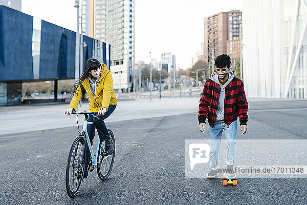 Lächelnde Frau auf dem Fahrrad neben einem Freund  der auf der Straße Skateboard fährt